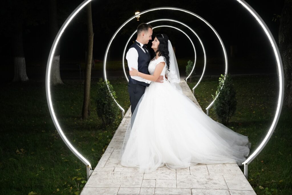 A couple embraces in wedding attire under illuminated rings at night, outdoors.