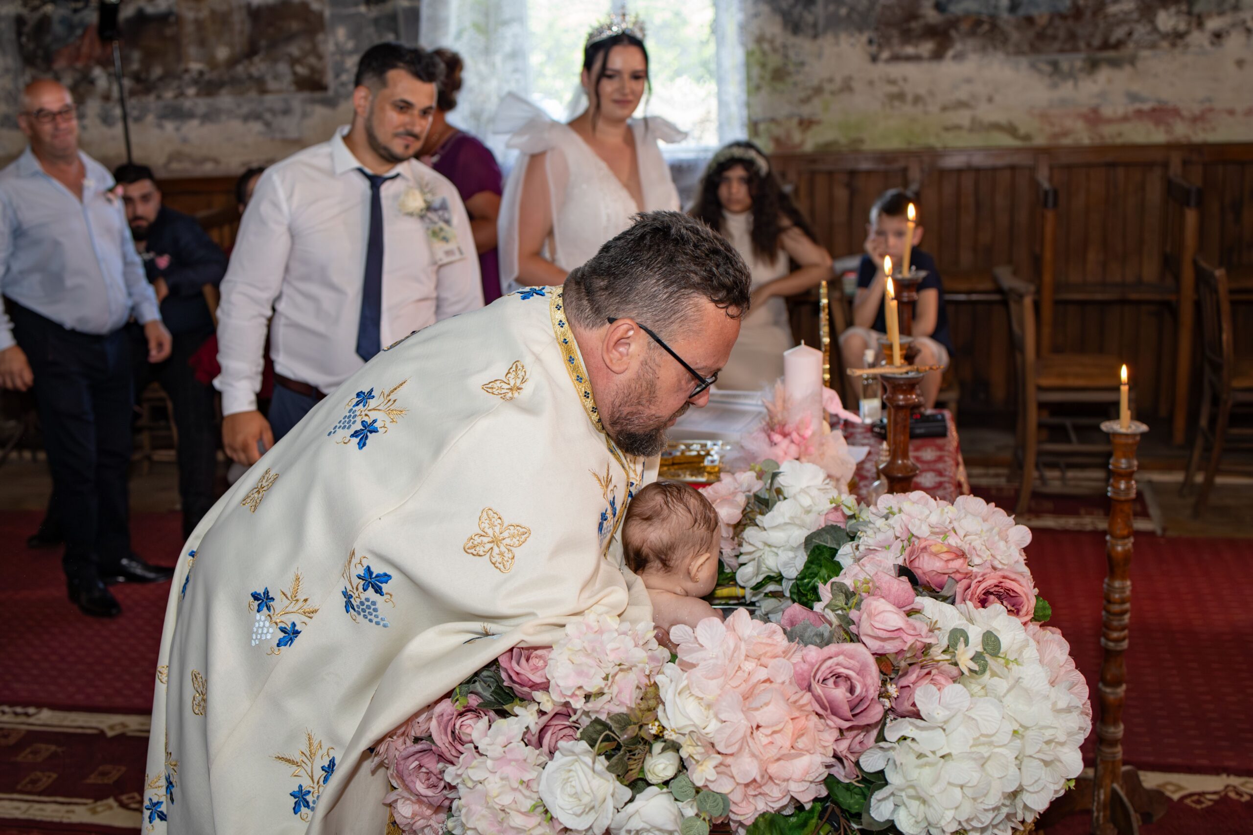 A traditional baptism ceremony with priest, parents, and guests in a historic church setting with floral decor.